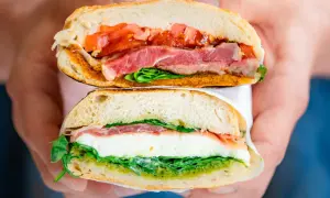 Deux moitiés d'un sandwich napolitain garni de jambon, mozzarella, tomates et pesto, tenues par des mains.