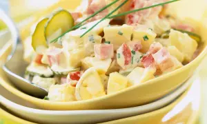 Salade piémontaise crémeuse avec pommes de terre, jambon, œufs durs, cornichons et ciboulette, servie dans un bol jaune.