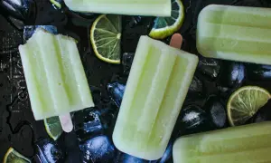 Plusieurs bâtonnets de glace au melon, avec des tranches de citron vert et des glaçons sur fond noir.