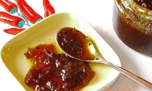 Une cuillère sert une délicieuse confiture de piment, avec un bocal et des piments rouges brodés en arrière-plan.