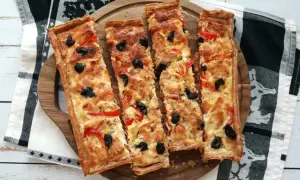 Quatre parts de tarte au thon dorée, garnie d'olives noires et de poivrons rouges, sur un plateau en bois.