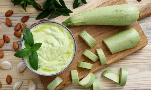 Un bol de purée de courgette crémeuse, garni de menthe fraîche, entouré d'amandes et d'ail. Des courgettes coupées en rondelles sur une planche à découper.