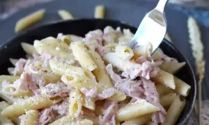 Une fourchette prélève des pâtes penne crémeuses avec du jambon effiloché, saupoudrées de parmesan et de poivre noir. Un plat de pâtes au jambon réconfortant.