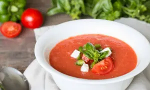 Un bol de gaspacho tomate frais garni de basilic, dés de poivrons et fromage. Des tomates, un poivron vert et du basilic frais en arrière-plan.