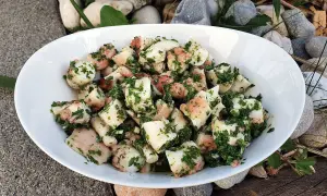 Salade de poulpe aux pommes de terre, assaisonnée d'herbes fraîches. Un délice méditerranéen servi dans un bol blanc, entouré de galets et de bois.