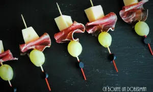 Délicieuses brochettes apéro : cubes de fromage, fines tranches de charcuterie et raisins verts sur fond noir.