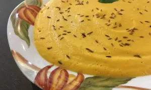 Velouté de carotte onctueux, parsemé de graines de cumin et garni d'une feuille de persil, servi dans une assiette décorée de motifs automnaux.