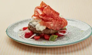 Burger garni de spianata piccante, burrata, tomates cerises et oignons rouges sur une assiette décorée.