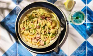 Un plat de pâtes aux coquillages et morceaux de poulpe, garni de persil frais et de quartiers de citron. Un verre de vin blanc et une fourchette complètent cette scène estivale.