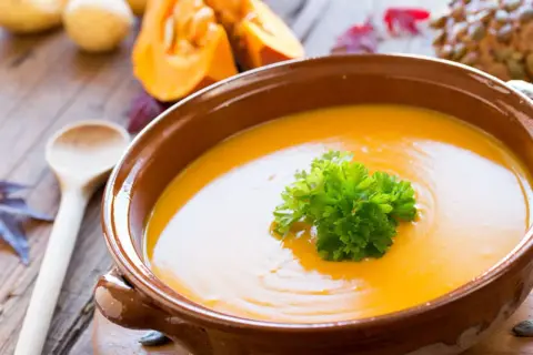 Soupe potimarron onctueuse, garnie de persil frais, dans un bol en terre cuite. Des morceaux de potimarron et une cuillère en bois complètent cette scène automnale.