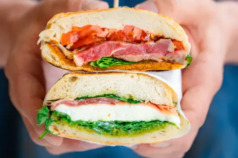 Deux moitiés d'un sandwich napolitain garni de jambon, mozzarella, tomates et pesto, tenues par des mains.
