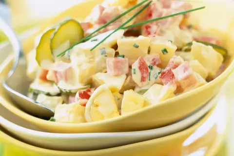 Salade piémontaise crémeuse avec pommes de terre, jambon, œufs durs, cornichons et ciboulette, servie dans un bol jaune.