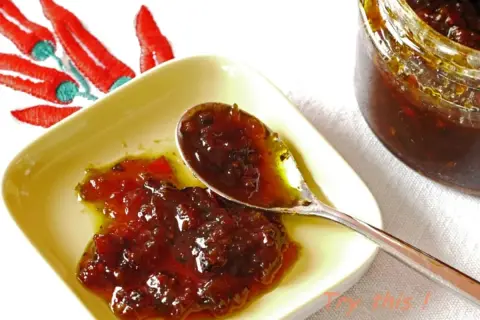 Une cuillère sert une délicieuse confiture de piment, avec un bocal et des piments rouges brodés en arrière-plan.