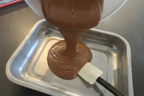 Une spatule aide à verser une ganache chocolat onctueuse dans un plat métallique.