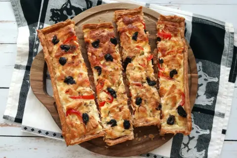 Quatre parts de tarte au thon dorée, garnie d'olives noires et de poivrons rouges, sur un plateau en bois.