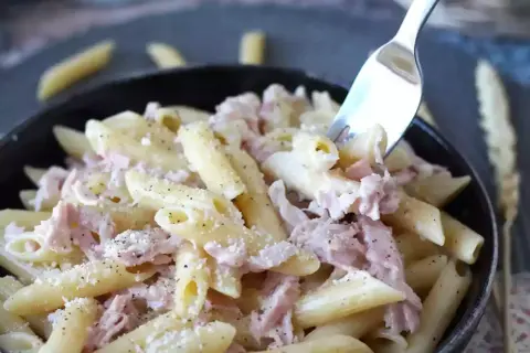 Une fourchette prélève des pâtes penne crémeuses avec du jambon effiloché, saupoudrées de parmesan et de poivre noir. Un plat de pâtes au jambon réconfortant.