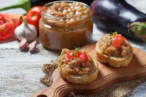 Deux toasts garnis de caviar d'aubergine, un pot de ce délice en arrière-plan, avec des aubergines, des tomates et de l'ail.