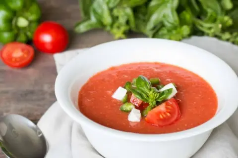 Un bol de gaspacho tomate frais garni de basilic, dés de poivrons et fromage. Des tomates, un poivron vert et du basilic frais en arrière-plan.