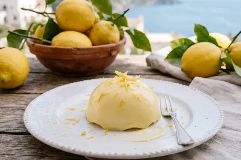 Un dessert crémeux, une **delizia al limone**, garni de zestes de citron, sur une assiette blanche, avec des citrons frais et la mer en arrière-plan.