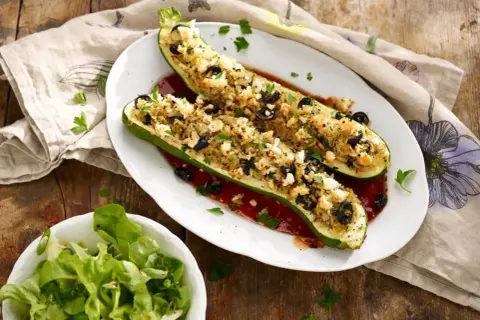 Deux courgettes farcies végétariennes, garnies de riz, olives et feta, sur un lit de sauce tomate. Une salade verte accompagne ce plat.