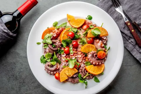 Salade de poulpe fraîchement préparée, garnie d'oranges, de tomates cerises et de verdure, prête à être dégustée avec une bouteille de vin rouge.
