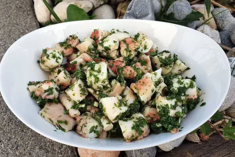 Salade de poulpe aux pommes de terre, assaisonnée d'herbes fraîches. Un délice méditerranéen servi dans un bol blanc, entouré de galets et de bois.
