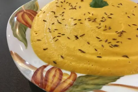 Velouté de carotte onctueux, parsemé de graines de cumin et garni d'une feuille de persil, servi dans une assiette décorée de motifs automnaux.