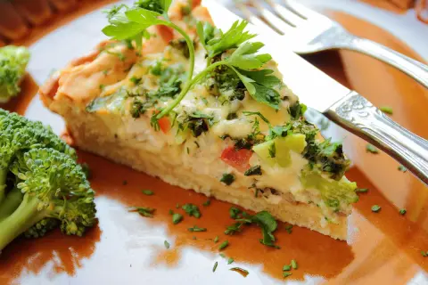 Une part de tarte aux légumes, garnie de persil frais, est servie sur une assiette orange avec du brocoli.