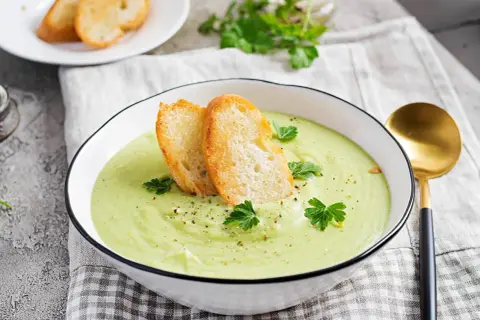 Un bol de soupe verte onctueuse, garnie de croûtons dorés et de persil frais. Parfait pour une recette gaspacho méditerranéen.