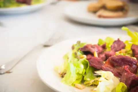 Salade périgourdine appétissante avec des morceaux de viande rouge, des pignons et de la laitue croquante sur une assiette blanche.