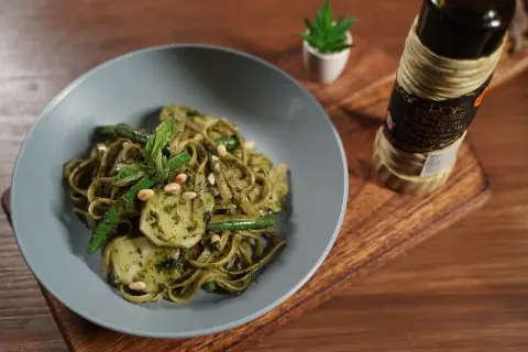 Assiette de trenette al pesto, garnie de haricots verts, pignons de pin et basilic frais. Une bouteille d'huile d'olive est visible à côté.