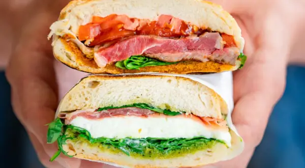 Deux moitiés d'un sandwich napolitain garni de jambon, mozzarella, tomates et pesto, tenues par des mains.