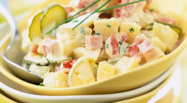 Salade piémontaise crémeuse avec pommes de terre, jambon, œufs durs, cornichons et ciboulette, servie dans un bol jaune.