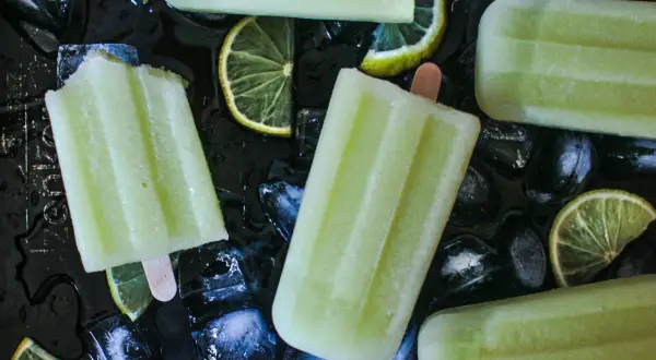 Plusieurs bâtonnets de glace au melon, avec des tranches de citron vert et des glaçons sur fond noir.