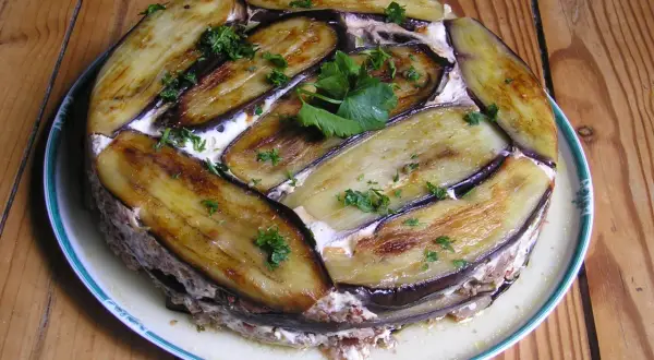 Moussaka ronde, une délicieuse moussaka ricetta, garnie d'aubergines grillées et de persil frais, sur une assiette blanche à bordure verte.