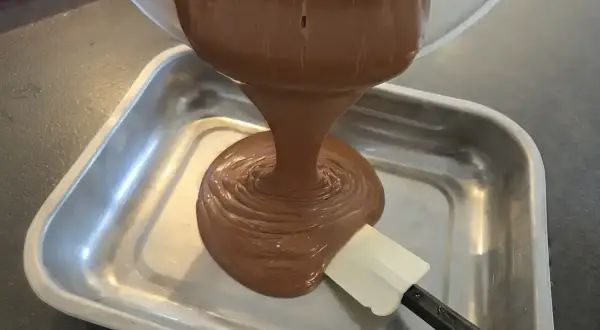 Une spatule aide à verser une ganache chocolat onctueuse dans un plat métallique.