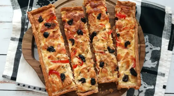 Quatre parts de tarte au thon dorée, garnie d'olives noires et de poivrons rouges, sur un plateau en bois.
