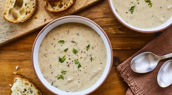 Deux bols de velouté de champignons à la crème, garni de persil et de poivre, avec des tranches de pain croustillant sur une planche en bois.