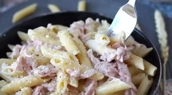 Une fourchette prélève des pâtes penne crémeuses avec du jambon effiloché, saupoudrées de parmesan et de poivre noir. Un plat de pâtes au jambon réconfortant.