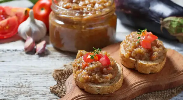 Deux toasts garnis de caviar d'aubergine, un pot de ce délice en arrière-plan, avec des aubergines, des tomates et de l'ail.
