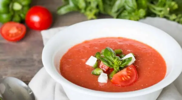 Un bol de gaspacho tomate frais garni de basilic, dés de poivrons et fromage. Des tomates, un poivron vert et du basilic frais en arrière-plan.