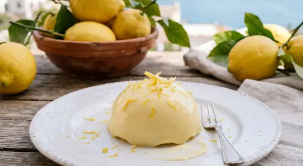 Un dessert crémeux, une **delizia al limone**, garni de zestes de citron, sur une assiette blanche, avec des citrons frais et la mer en arrière-plan.