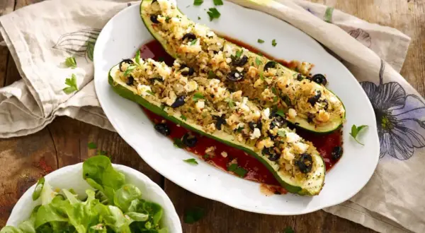 Deux courgettes farcies végétariennes, garnies de riz, olives et feta, sur un lit de sauce tomate. Une salade verte accompagne ce plat.