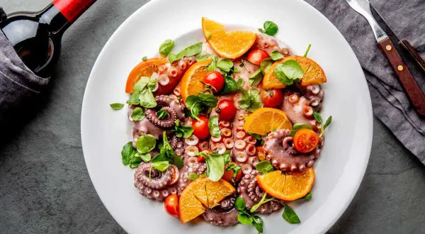 Salade de poulpe fraîchement préparée, garnie d'oranges, de tomates cerises et de verdure, prête à être dégustée avec une bouteille de vin rouge.