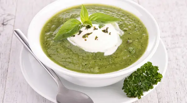 Soupe froide courgette fromage frais, garnie de basilic et persil. Une cuillère repose à côté du bol.