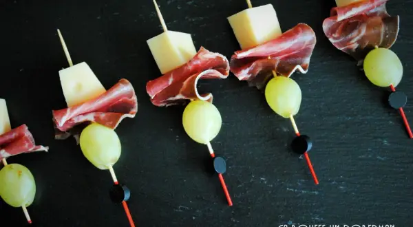 Délicieuses brochettes apéro : cubes de fromage, fines tranches de charcuterie et raisins verts sur fond noir.