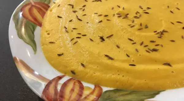 Velouté de carotte onctueux, parsemé de graines de cumin et garni d'une feuille de persil, servi dans une assiette décorée de motifs automnaux.