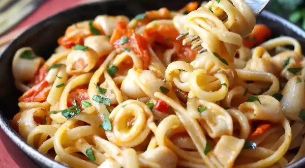Une assiette de pâtes aux calamars italienne, garnie de persil frais et de morceaux de tomates. Une fourchette soulève une portion de ce plat savoureux.