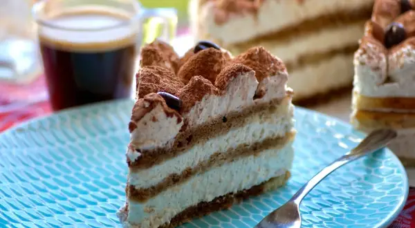 Une part de tiramisu gâteau, crémeux et aérien, saupoudrée de cacao, avec un café noir en arrière-plan.