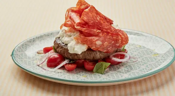 Burger garni de spianata piccante, burrata, tomates cerises et oignons rouges sur une assiette décorée.